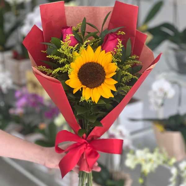 Pessoa segurando o Buquê Camélia Brilho e Paixão, mostrando o tamanho real do girassol com 2 rosas vermelhas e a embalagem moderna com dobraduras da Floricultura Camélia em São Paulo Zona Norte.